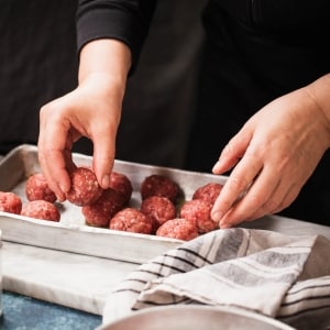 Las manos de un chef colocan albóndigas de carne de res recién preparadas en una bandeja de horno. En la mesada también se ven un trapo de cocina y condimentos.