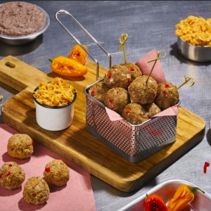 Albóndigas vegetarianas de frijol servidas en formato brochetas en una canasta metálica con un papel rosado. Sobre la tabla en la que se apoya, también hay un ramekin con kimchi y pimientos naranjas. En la mesada de acero inoxidable también se ven contenedores con puré de frijoles refritos, kimchi y más pimientos, y también hay más albóndigas sobre otro papel.