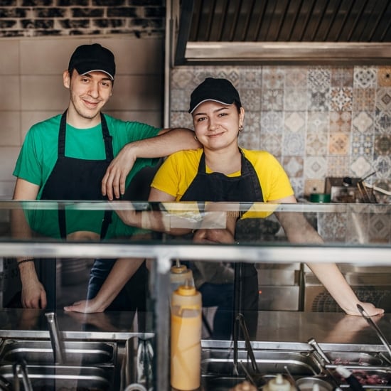 Dos empleados de un negocio de comida sonríen. Él apoya su codo en el hombro de ella. Delante de ellos se ve el mostrador de despacho, con bateas con diferentes ingredientes y botellas dispensadoras de aderezos.