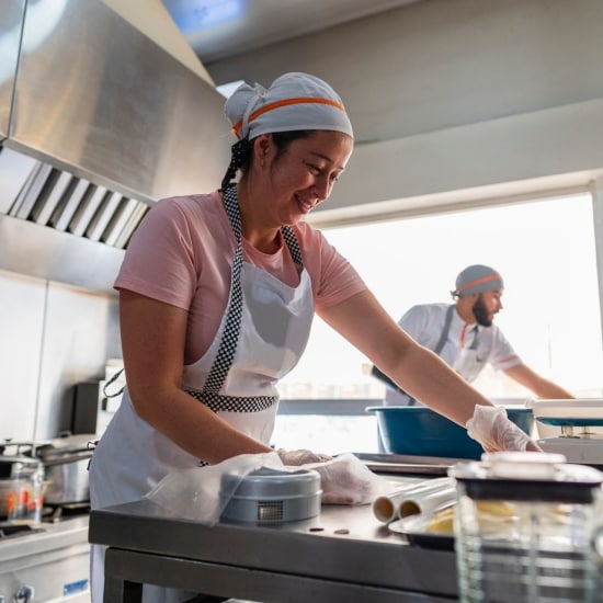 Dos cocineros trabajan en una cocina comercial. En primer plano, la mujer empaqueta pedidos.