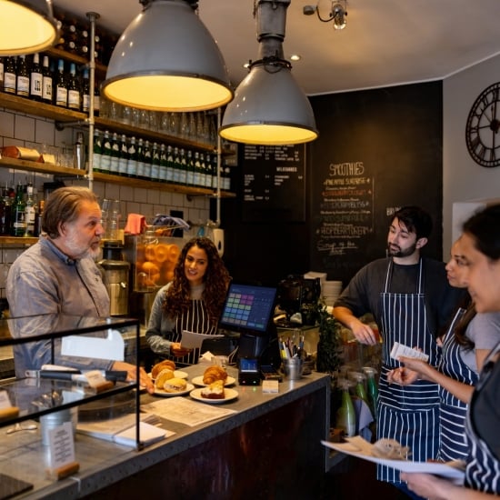El dueño de una cafetería conversa con cinco empleados que lo miran atentamente y sonríen mientras toman notas. Están de pie alrededor de la barra, en donde hay platos con panes dulces, bollería y pastelería.