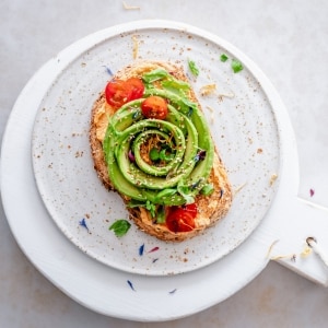 Pan blanco tostado con una capa de humus con paprika, tomates uva rojos cortados a la mitad y aguacate cortado en rodajas y servido con forma de flor. Está servido sobre un plato blanco, apoyado en una tabla blanca, y decorado con hojas de albahaca.