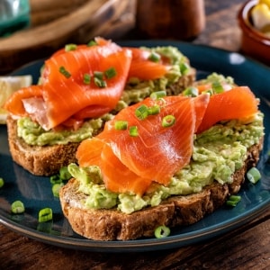 Dos avocado toast con pan integral, aguacate machacado y fetas de salmón gravlax, decorado con cebollín picado. Están servidos en platos azules sobre mesa de madera.