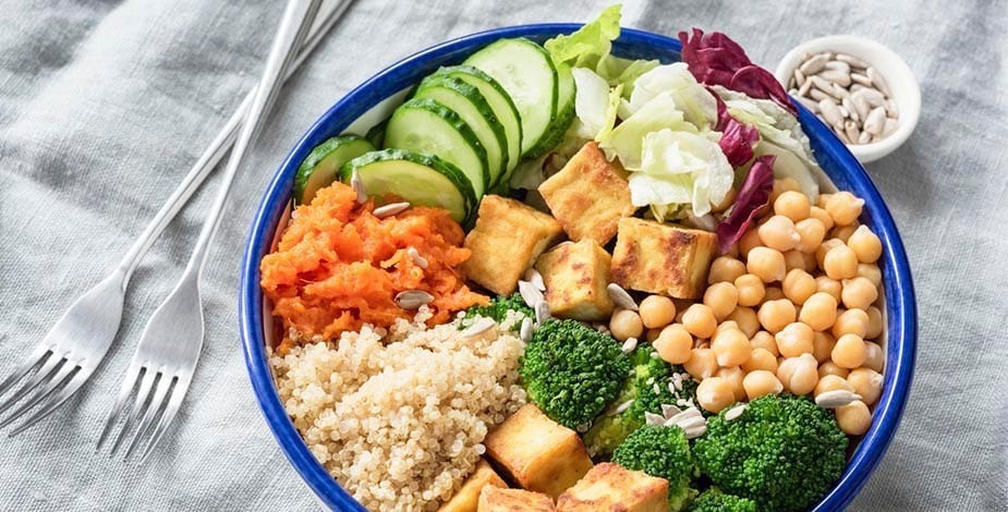 bowl, buddha bowl, con quinoa, garbanzos, tofu, brócoli, zanahoria, lechuga, pepino sobre mantel claro y acompañado de semillas de girasol