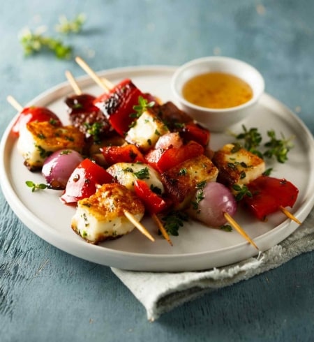 Brochetas de queso, cebolla morada y pimiento rojo asadas a la parrilla, servidas con hierbas y con una salsa en un plato blanco. Brochetas de queso, cebolla morada y pimiento rojo asadas a la parrilla, servidas con hierbas y con una salsa en un plato blanco.