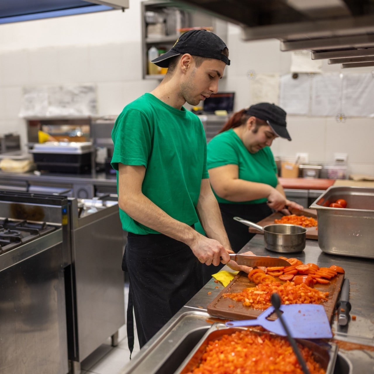 Dos cocineros pican jitomate parados uno al lado del otro en la estación de cocina, utilizando tablas sobre las mesas de acero inoxidable. Dos cocineros pican jitomate parados uno al lado del otro en la estación de cocina, utilizando tablas sobre las mesas de acero inoxidable.