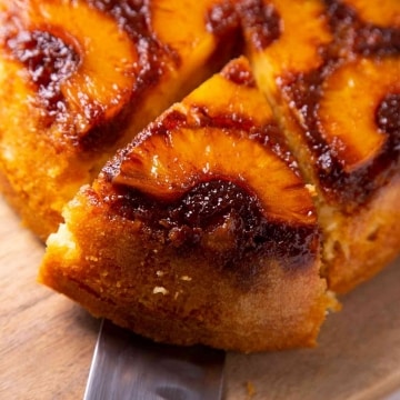 Una persona toma una porción de pastel de piña invertido utilizando una espátula. El pastel es de bizcocho y tiene una capa de piña caramelizada