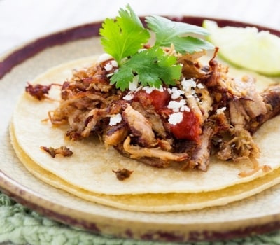 Tacos de carnitas, con carne de cerdo deshebrada, salsa roja, queso y cilantro frescos servidos con dos tortillas de maíz y una rodaja de limón.