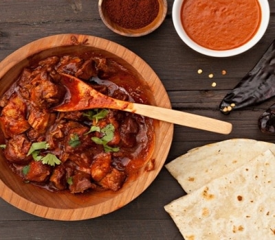 Carne de cerdo estofado en salsa de chili rojo, servido en un plato de madera y acompañado por tortillas de harina.