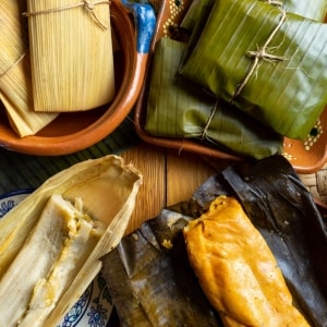 Variedad de tamales, algunos envueltos en hoja de maíz de color pajizo y en hoja de plátano de tonalidad verde oscura, y otros abiertos, en los que se ve las hojas desplegadas y la masa y el relleno ya cocidos.