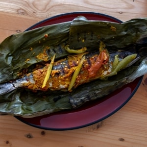 Pescado entero servido sobre hojas de plátano en las que estuvo envuelto durante la cocción. También se ven trozos de jitomate y pimiento verde. Está servido en un plato púrpura, sobre una mesa de madera.