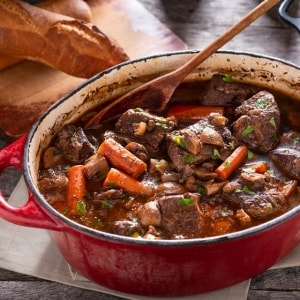 Estofado de carne de res con caldo, zanahorias, champiñones y hojas de perejil en una olla de hierro fundido color rojo. Estofado de carne de res con caldo, zanahorias, champiñones y hojas de perejil en una olla de hierro fundido color rojo.