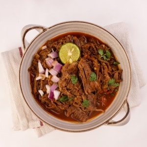 Birria, platillo típico mexicano de carne de res preparada en cocción lenta, servida en una fuente de cerámica junto a una rodaja de limón verde, cebolla morada picada y hojas de cilantro. Birria, platillo típico mexicano de carne de res preparada en cocción lenta, servida en una fuente de cerámica junto a una rodaja de limón verde, cebolla morada picada y hojas de cilantro.