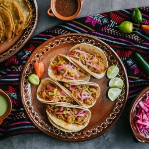 Cinco tacos de cochinita pibil servidos con cebolla encurtida y tajadas de limón verde en un plato tradicional mexicano. Acompañan otros contenedores típicos con salsa verde, salsa roja y cebolla encurtida, y otro platillo preparado a base de tortillas crujientes. De fondo se ve un mantel colorido local, y sobre él diferentes tipos de chiles.