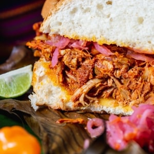 Torta de cochinita pibil preparada con pan esponjoso y relleno de cerdo deshebrado con cebollas encurtidas. Sobre la tabla también hay limón verde, chiles y más cebolla.