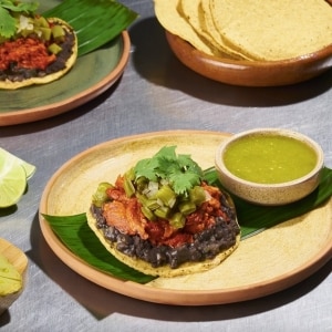 Tostada con base de maíz, frijoles refritos, cochinita pibil, nopales en conserva y hojas de cilantro, servida sobre una hoja de plátano en un plato beige que también tiene un pote de salsa verde. Sobre la mesada de acero inoxidable también se ven otra tostada y tortillas.