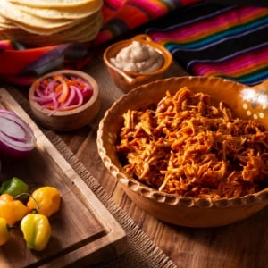 Cochinita pibil deshebrada en un plato de barro mexicano. En la mesa también hay una tabla con cebolla y chiles crudos, dos potes con cebolla encurtida y una salsa, tortillas y un mantel colorido.