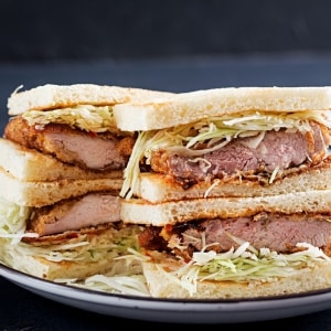 Cuatro porciones de katsu sando, un sándwich típico japonés preparado con lomo de cerdo empanado, col blanca y salsa agridulce en pan tierno de miga. Están servidos de a dos, uno arriba del otro, sobre un planto blando de borde azul.