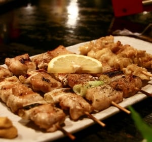 Plato de yakitori, brochetas típicas de la comida japonesa. Están hechas con trozos de pollo y cerdo marinados, y asados en la parrilla junto a cebollín. Servidas en una fuente blanca, con media rodaja de limón amarillo arriba.