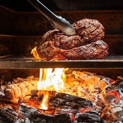 Close-up view of a perfectly grilled steak being flipped over burning wood on an open flame. The glowing embers and dancing flames add warmth and authenticity, capturing the essence of outdoor cooking and smoky flavors.