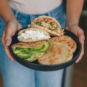 Una mujer sostiene con sus dos manos un plato con cinco arepas, dos rellenas y dos simples. Entre las rellenas, hay de pollo, de queso fresco y de aguacate. La mujer viste jeans azules y playera blanca.