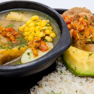 Plato sopero con sancocho, una sopa con caldo, maíz, pollo, jitomate y papa, servido junto a una pierna de pollo, una rebanada de aguacate y arroz blanco.
