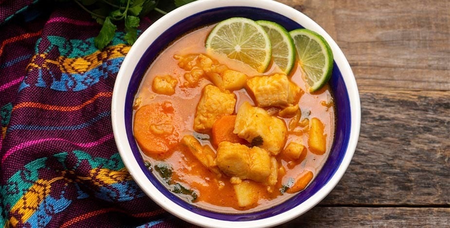 sopa de pescado con vegetales y rodajas de lima en plato de cerámica blanco y azul sobre mesa de madera con mantel tejido típico mexicano