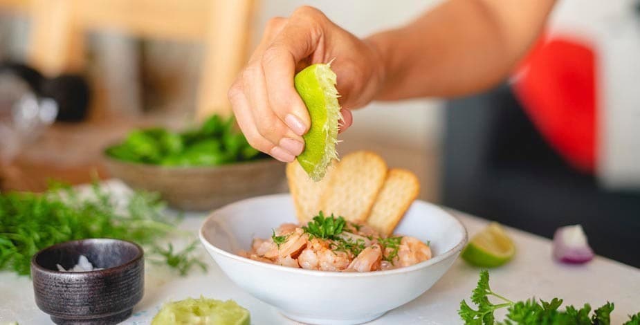 mano de mujer exprimiendo una lima sobre un ceviche de camarones, sobre mesada blanca con sal marina, cilantro y lima