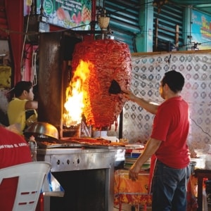 Maestro taquero prepara carne de pastor en un trompo, usando una espátula para repasar la carne. También se ven la llama que sale del horno pastorero, y a otros empleados de la taquería.