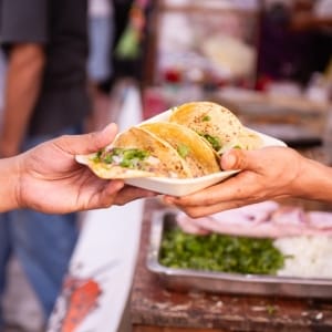 La mano de un vendedor de comida callejera mexicana entrega un plato con tres tacos a un cliente. De fondo se ve una bandeja con cebolla picada y hojas de cilantro.
