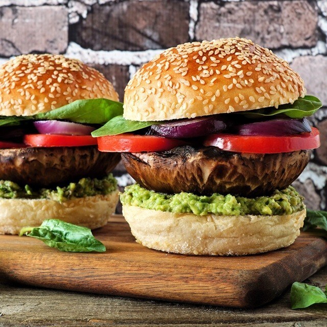 Hamburguesas veganas con pan con sésamo, portobello, tomate, cebolla, aguacate y hojas verdes, servidas sobre una tabla. Hamburguesas veganas con pan con sésamo, portobello, tomate, cebolla, aguacate y hojas verdes, servidas sobre una tabla.