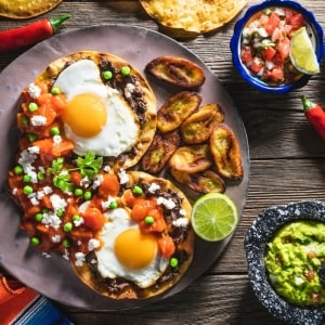 Huevos motuleños, platillo compuesto por una tortilla frita untada con frijoles refritos, encima el huevo estrellado, jitomate cocido, chícharos y queso fresco, acompañados con plátanos machos fritos. En la mesa también hay contenedores con guacamole y pico de gallo, y tajadas de limón verde