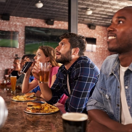 Un grupo de amigos mira atentamente un partido de fútbol en la barra de un bar mientras toman cerveza y refrescos y comen hamburguesas con papas a la francesa. De fondo se ven más pantallas con la transmisión.