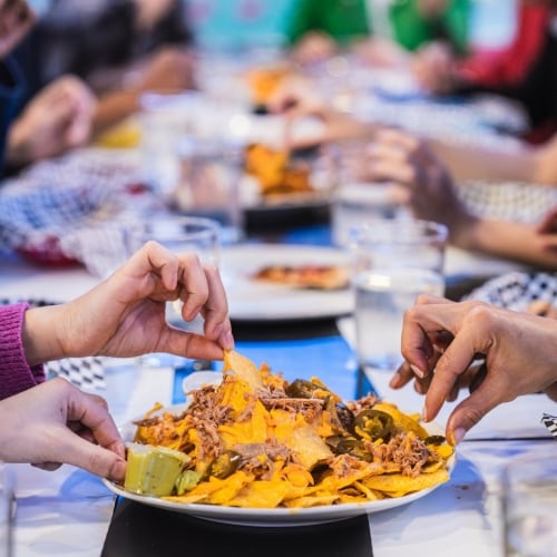 En una mesa larga, un grupo de amigos comparte varios platos de comida, entre ellos uno de nachos con jalapeños, carne desmechada y guacamole. Los comensales agarran las tortillas fritas con las manos. En la mesa también hay vasos de agua.