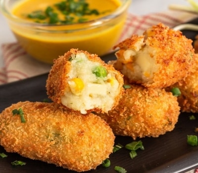 Croquetas alargadas de chícharos, maíz y queso, con interior cremoso y empanizado crocante y dorado. Están servidas con un aderezo de mostaza y con cebollín fresco por arriba.