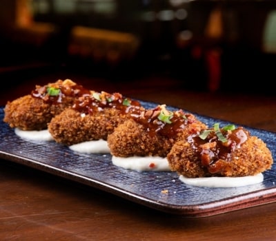 Cuatro croquetas doradas servidas sobre una salsa blanca en un plato azul. Arriba llevan una salsa de tomate con tocino crocante y cebollín.