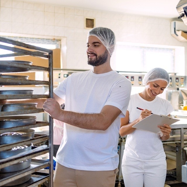 Dos cocineros de una panadería trabajan juntos. Mientras ella controla el stock de ingredientes, él acomoda fuentes en un rack.