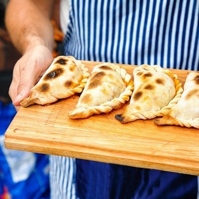 Close-up on perfectly baked empanadas tucumanas on a cutting board held by baker outdoors.