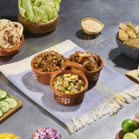 Tres guisados para preparar tacos servidos en cuencos de barro, sobre mantel azul, rodeado de tortillas, verduras y frutas. Tres guisados para preparar tacos servidos en cuencos de barro, sobre mantel azul, rodeado de tortillas, verduras y frutas.