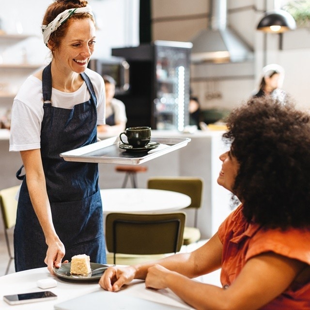 Mesera amable sirve un café y un pastel en la mesa a una clienta sonriente, mientras conversan en la cafetería. Mesera amable sirve un café y un pastel en la mesa a una clienta sonriente, mientras conversan en la cafetería.