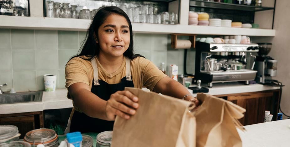 mujer hispana en mostrador de cafetería o restaurante entregando dos paquetes para delivery