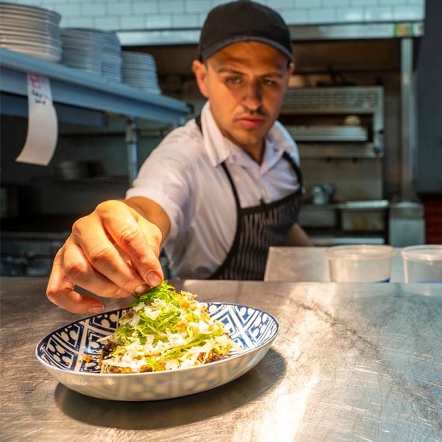 chef latino trabajando en cocina, terminando de emplatar una ensalada en mostrador de acero