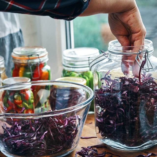 Hombre prepara alimentos fermentados, colocando col morada cortada en un recipiente de vidrio. Atrás se ven otros ingredientes fermentados. Hombre prepara alimentos fermentados, colocando col morada cortada en un recipiente de vidrio. Atrás se ven otros ingredientes fermentados.