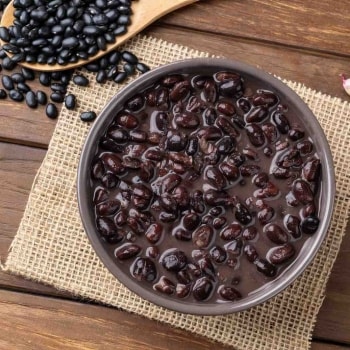Frijoles negros guisados servidos en una fuente gris que está apoyada sobre un paño de fibras. Sobre la mesa de madera también hay una cuchara con frijoles negros crudos. Frijoles negros guisados servidos en una fuente gris que está apoyada sobre un paño de fibras. Sobre la mesa de madera también hay una cuchara con frijoles negros crudos.