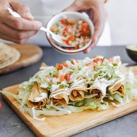 cocinero añadiendo salsa de tomate y cebolla a unas flautas de pollo servidas sobre hojas de lechuga en tabla de madera