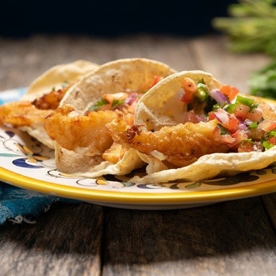 Tacos de pescado frito, conocidos como tacos ensenada, servidos en tortillas de trigo con una salsa de jitomate, chiles y cebolla cruda cortados en cubos. 