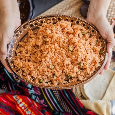 Las manos de una cocinera ofrecen arroz rojo con chícharos y zanahorias. Está servido en un plato de barro tradicional, y de fondo también se ven manteles de estilo mexicano. Las manos de una cocinera ofrecen arroz rojo con chícharos y zanahorias. Está servido en un plato de barro tradicional, y de fondo también se ven manteles de estilo mexicano.