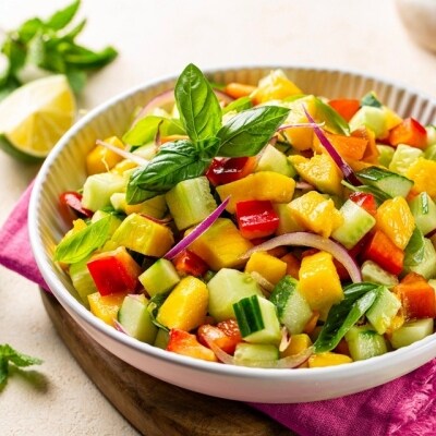 Ensalada de pepino, jitomate, mango, pimiento y albahaca, servida en un bol de cerámica blanco, apoyada sobre un paño rosa y una tabla. De fondo se ven tajadas de limón y más hojas. Ensalada de pepino, jitomate, mango, pimiento y albahaca, servida en un bol de cerámica blanco, apoyada sobre un paño rosa y una tabla. De fondo se ven tajadas de limón y más hojas.