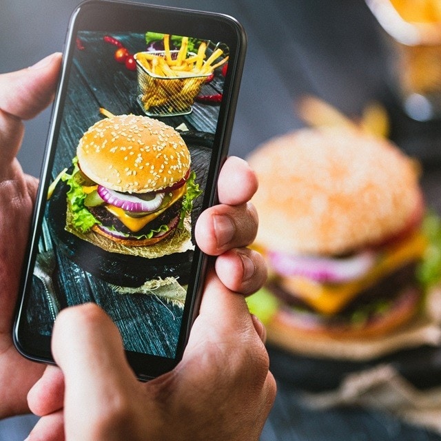 Persona saca una foto con el teléfono celular de una hamburguesa de carne con pan, lechuga, queso y cebolla, junto a una canasta de papas a la francesa. Persona saca una foto con el teléfono celular de una hamburguesa de carne con pan, lechuga, queso y cebolla, junto a una canasta de papas a la francesa.