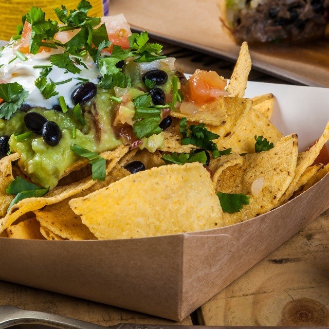 Tortillas de maíz horneadas servidas en un empaque de cartón sostenible, acompañadas de frijoles negros, guacamole, pico de gallo y cilantro. Tortillas de maíz horneadas servidas en un empaque de cartón sostenible, acompañadas de frijoles negros, guacamole, pico de gallo y cilantro.